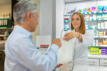 Positive junge weibliche Kassiererin mit blondem Haar, die einem älteren männlichen Kunden eine Papiertüte gibt, während sie an der Kasse eines Pharmaziegeschäfts steht - ADSF50373
