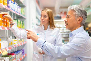 Young female pharmacist in white uniform assisting senior male customer in buying and choosing drugs from shelves at store - ADSF50375