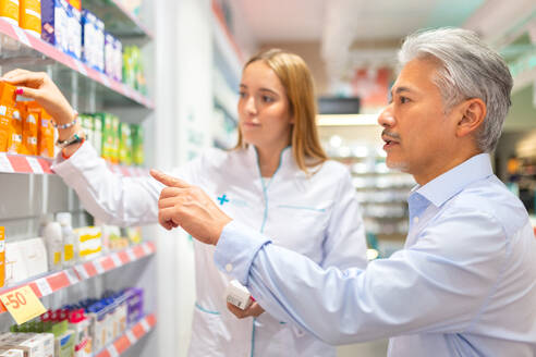 Junge Apothekerin in weißer Uniform, die einem älteren männlichen Kunden beim Kauf und bei der Auswahl von Medikamenten aus den Regalen eines Geschäfts hilft - ADSF50375