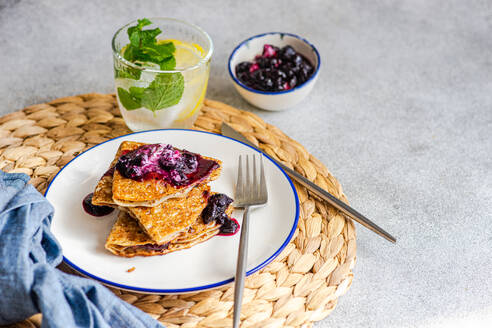 Ein Stapel frischer, hausgemachter Crêpes, belegt mit Blaubeermarmelade und einem Klecks saurer Sahne und Zitronenwasser, präsentiert auf einem weißen Teller mit einem Tischset aus Weide. - ADSF50386