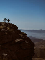 Silhouetted figures of two anonymous cyclists carrying their bikes up a steep mountain cliff at dusk - ADSF50426