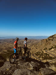 Seitenansicht zwei Radfahrer stehen mit ihren Mountainbikes mit Blick auf eine kurvenreiche Straße und sanften Hügeln - ADSF50430