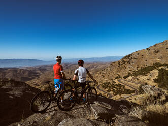 Blick zurück auf zwei Radfahrer, die mit ihren Mountainbikes stehen und eine kurvenreiche Straße und sanfte Hügel überblicken - ADSF50431