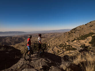 Seitenansicht: Zwei Mountainbiker machen eine Pause, um die Aussicht auf eine sich durch die Hügel schlängelnde Serpentinenstraße zu genießen - ADSF50432