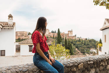 Seitenansicht einer anonymen jungen Touristin, die auf einer Stützmauer sitzt und auf die historische Alcazaba und Bäume gegen den bewölkten Himmel in Granada, Spanien, blickt - ADSF50446
