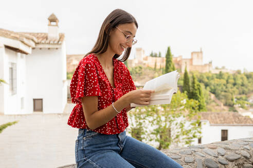 Glückliche, konzentrierte junge Frau in Freizeitkleidung und mit Brille, die ein Buch liest, während sie auf einer Stützmauer mit der Festung Alcazaba und dem klaren Himmel im Hintergrund sitzt - ADSF50447