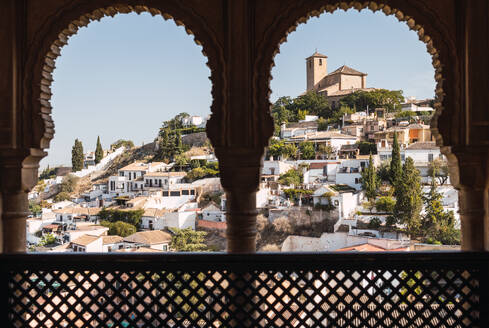 Schöner Blick auf das Stadtbild mit Glockenturm und Wohngebäuden auf einem Hügel gegen den klaren Himmel, gesehen durch die Bogenfenster des Generalife-Palastes in Granada, Spanien - ADSF50449