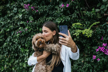 Young woman kissing and taking selfie with poodle dog in front of plants - GDBF00118