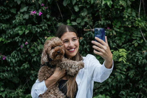 Happy woman taking selfie with poodle dog in front of plants - GDBF00119