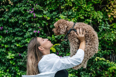 Happy young woman playing with pet poodle dog in front of plants - GDBF00121