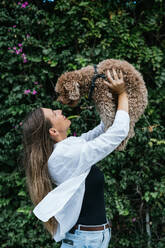 Happy young woman holding poodle dog aloft in front of plants - GDBF00122