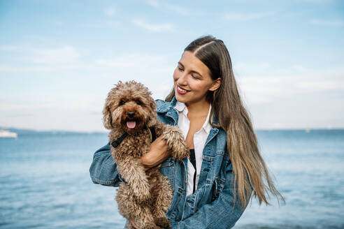 Smiling woman with poodle dog in front of sea - GDBF00129