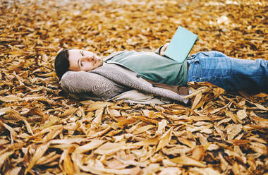 Young man holding book and lying on autumn leaves - SVCF00431
