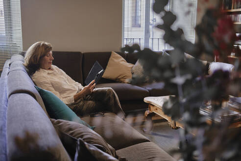 Mature woman sitting on sofa and using tablet PC at home - MMPF01085