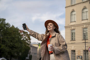 Happy woman holding smart phone and hailing ride near building - VPIF09035