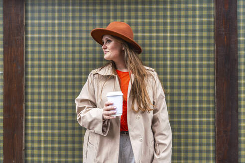 Lächelnde Frau mit Hut und Kaffeetasse in der Hand vor einer karierten Wand - VPIF09037