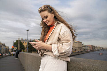 Smiling woman using smart phone near wall under cloudy sky in city - VPIF09047