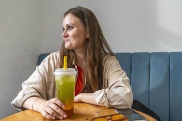 Smiling woman holding glass of juice and sitting in cafe - VPIF09050