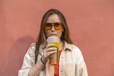 Young woman drinking juice in front of peach colored wall - VPIF09051