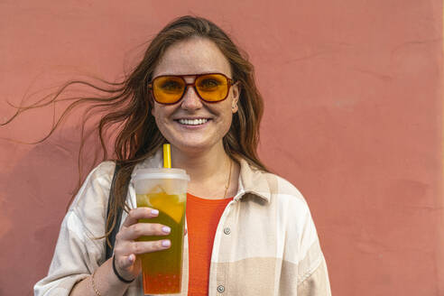 Happy young woman holding juice in front of peach colored wall - VPIF09052