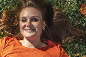 Glückliche rothaarige Frau mit Sonnenlicht über dem Gesicht, die sich im Gras entspannt - VPIF09077