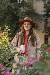 Lächelnde junge Frau mit Hut und Kaffeetasse in der Nähe von blühenden Pflanzen - VPIF09084