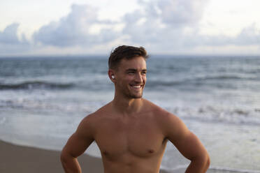Happy shirtless man wearing wireless in-ear headphones at beach - STF00032