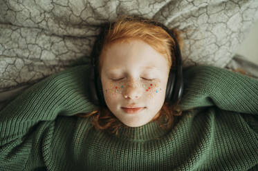 Redhead girl listening to music through wireless headphones at home - ANAF02587