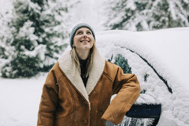 Woman wearing winter coat and leaning on car in snow - VSNF01499
