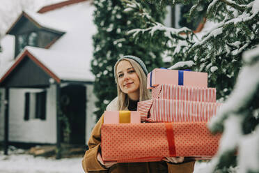Smiling woman holding Christmas presents near tree - VSNF01514