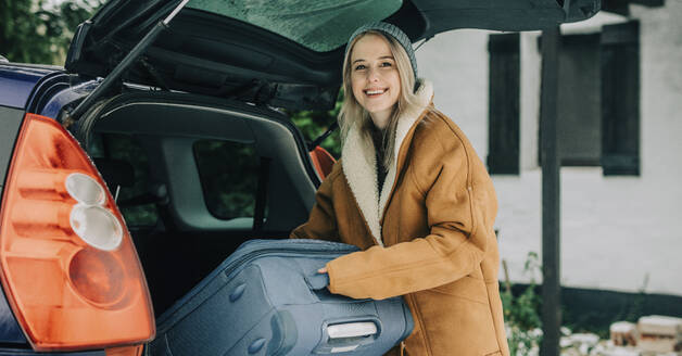 Happy woman putting suitcase in car trunk - VSNF01525