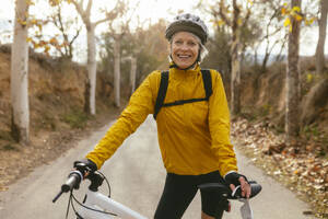 Smiling woman standing with mountain bike on road in forest - EBSF04300