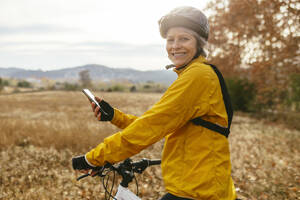 Smiling woman sitting on mountain bike with smart phone in field - EBSF04302