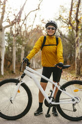 Smiling woman standing with mountain bike on road in forest - EBSF04312