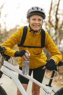 Smiling woman holding water bottle and leaning on mountain bike in forest - EBSF04314