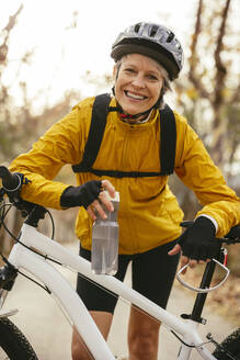 Happy woman holding water bottle and leaning on mountain bike in forest - EBSF04315