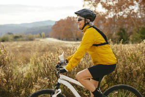 Happy woman riding mountain bike near field - EBSF04316
