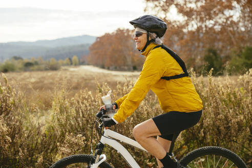 Happy woman riding mountain bike near field - EBSF04316
