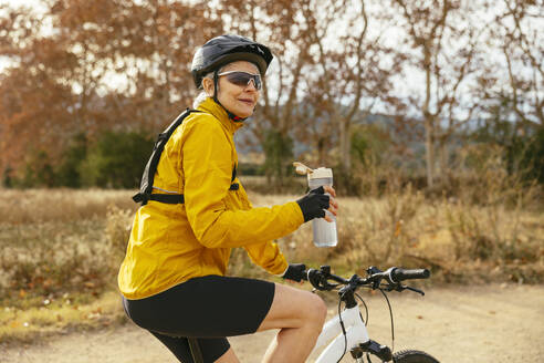Happy woman sitting on mountain bike with water bottle near field - EBSF04317