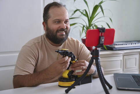Lächelnder Ingenieur, der zu Hause eine Präsentation hält und einen Vlog über einen Roboterarm auf seinem Smartphone führt - AZF00590