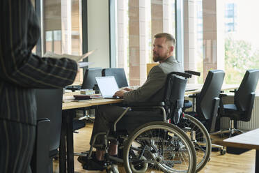 Geschäftsfrau im Gespräch mit einem Mitarbeiter, der im Rollstuhl sitzt und einen Laptop am Schreibtisch im Büro hat - DSHF01459
