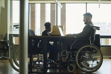 Businessman in wheelchair working on laptop with coworker in office - DSHF01460