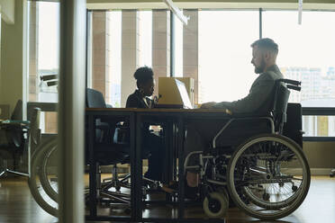 Geschäftsmann im Rollstuhl arbeitet am Laptop mit Mitarbeiter im Büro - DSHF01460