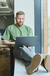 Lächelnder Geschäftsmann sitzt auf der Fensterbank und benutzt einen Laptop im Büro - DSHF01470