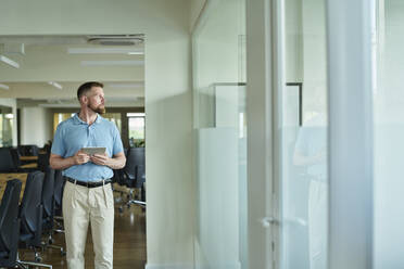 Contemplative businessman holding tablet PC and standing in office - DSHF01485