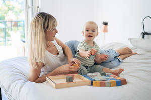 Mother and son playing with toy blocks on bed at home - JOSEF22385