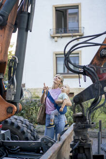 Mother carrying son and pointing at bulldozer near building - JOSEF22393