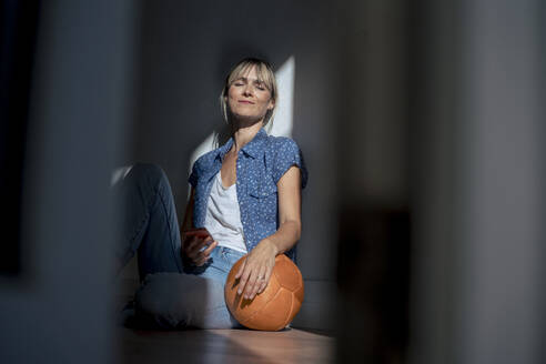 Woman with eyes closed sitting with ball near wall at home - JOSEF22411