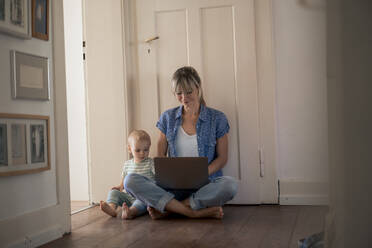 Smiling mother using laptop and sitting with son near door at home - JOSEF22416