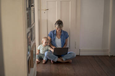 Working mother using laptop with son sitting on floor at home - JOSEF22417